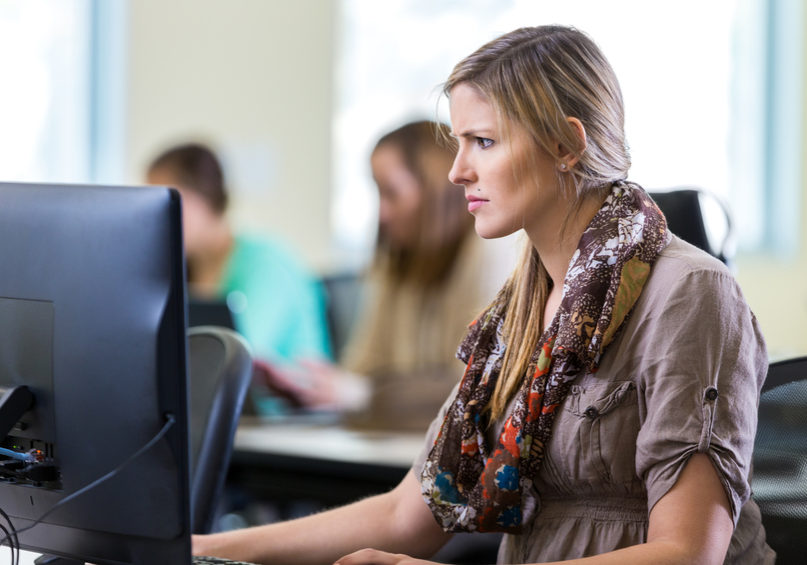 Mid adult Caucasian woman has an angry expression while looking at desktop computer monitor. She is sitting at a desk in a computer lab in public library. Woman is wearing casual clothing and a printed scarf.